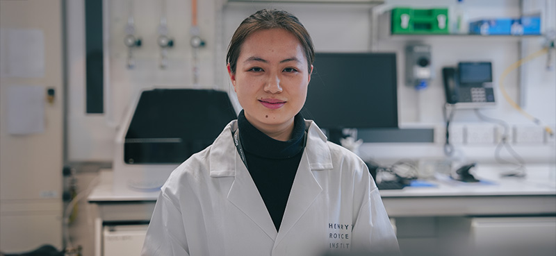 Researcher in a laboratory wearing a white lab coat, standing in front of scientific equipment.
